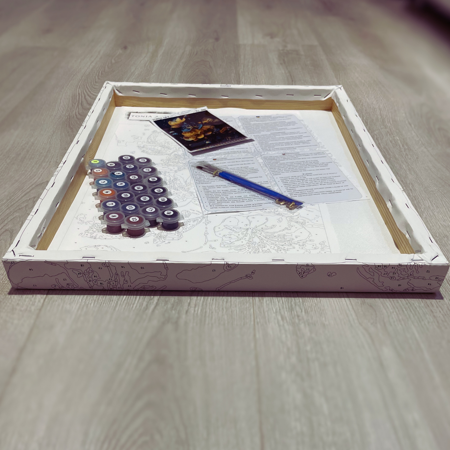 Un kit de peinture par numéros disposé sur le dos d’une toile encadrée, posée sur un sol en bois. Le kit comprend des pots de peinture numérotés, un pinceau bleu, des instructions et une image de référence de l'œuvre finie