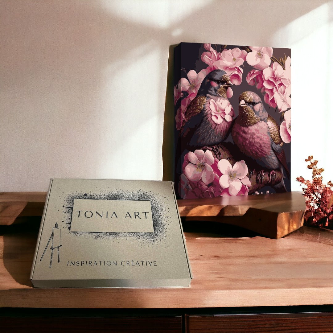 Le tableau représente deux colombes dans des fleurs roses. Le tableau est placé sur un podium en bois, à côté d'une boîte en carton portant l'inscription Tonia Art.