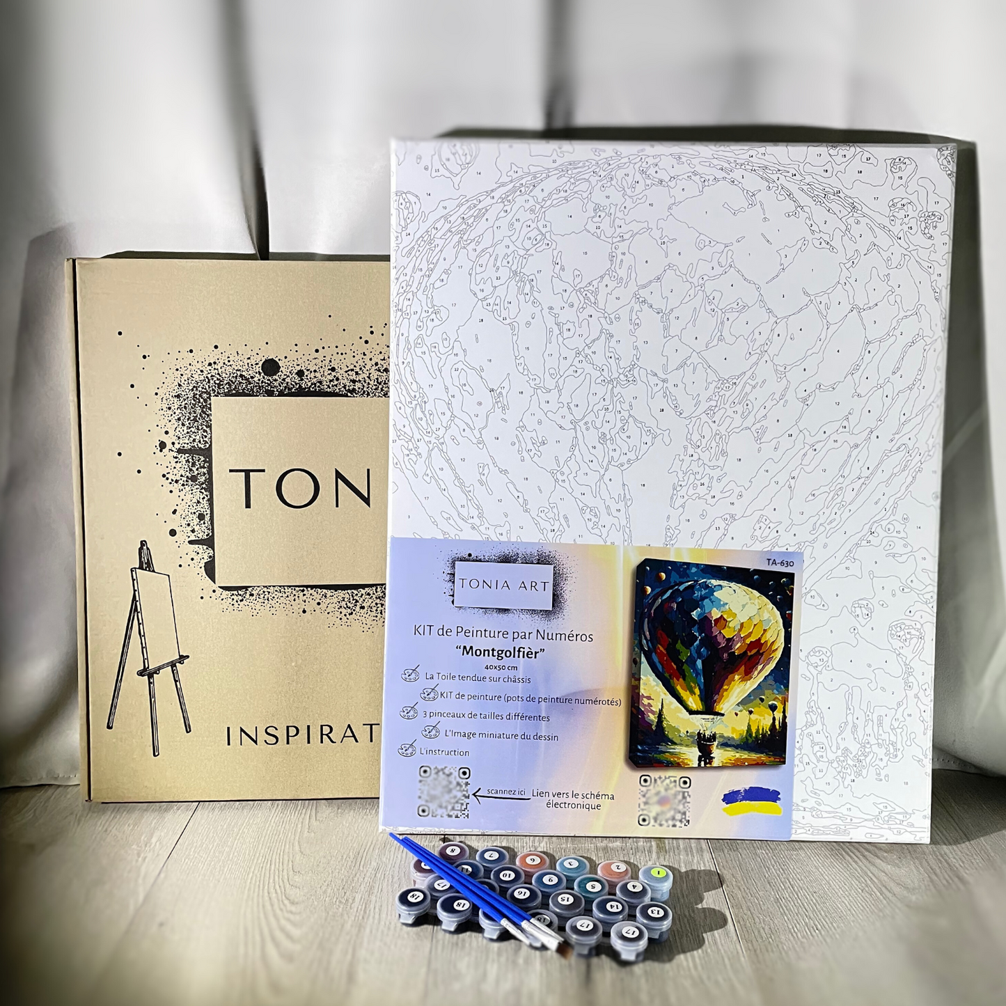 Kit de peinture par numéros de la marque Tonia Art, intitulé "Montgolfier," comprenant une toile imprimée avec le dessin d'une montgolfière, des pots de peinture numérotés, trois pinceaux de différentes tailles, et une boîte en carton marron avec le logo de la marque. Une petite image de référence colorée montre le résultat final.