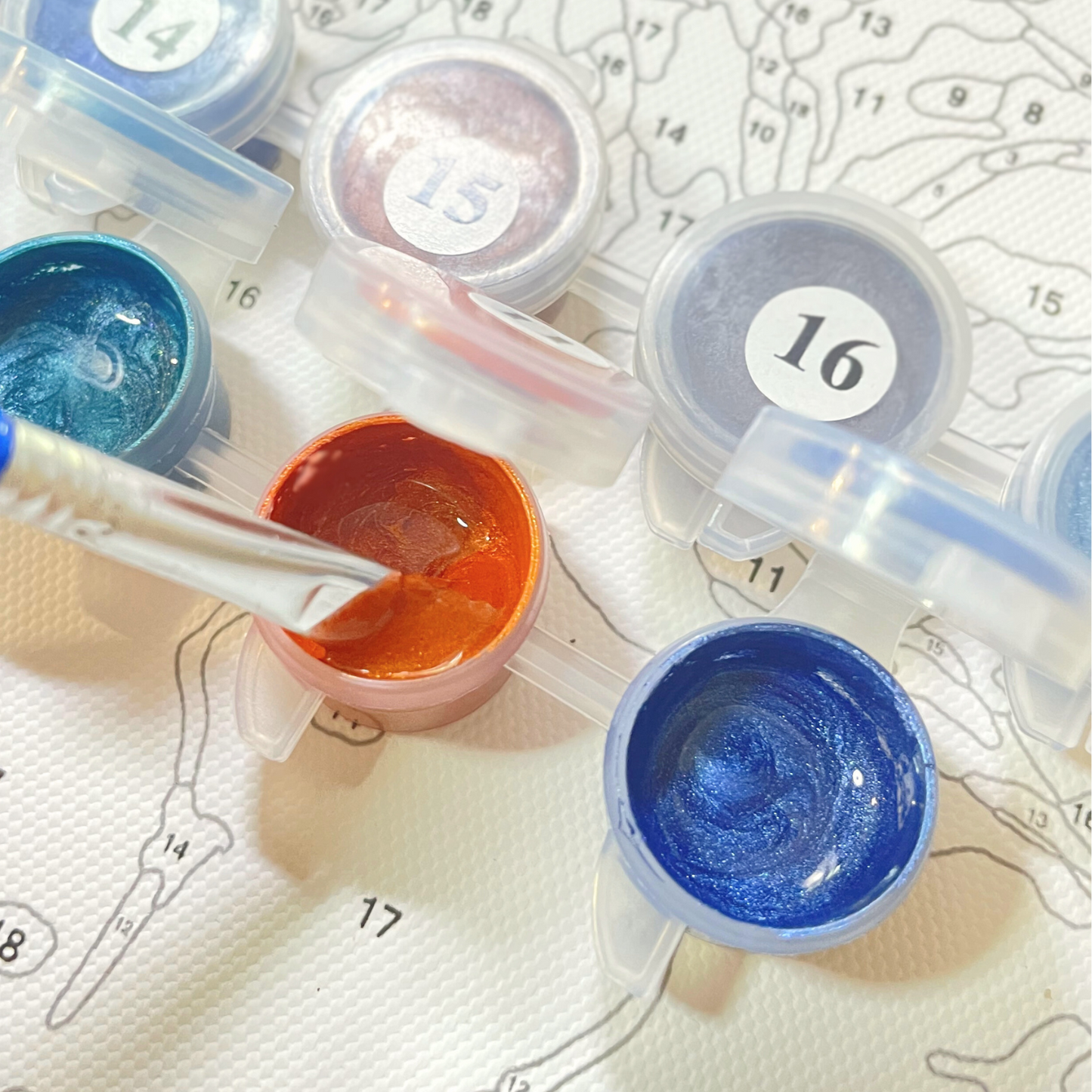 Gros plan d’un kit de peinture par numéros avec de petits pots de peinture aux nuances métalliques. Chaque pot est numéroté pour correspondre aux sections de la toile en dessous. Un pinceau est en train de plonger dans un pot de peinture orange vif, avec d'autres couleurs comme le bleu sarcelle, le bleu et le lavande à proximité.