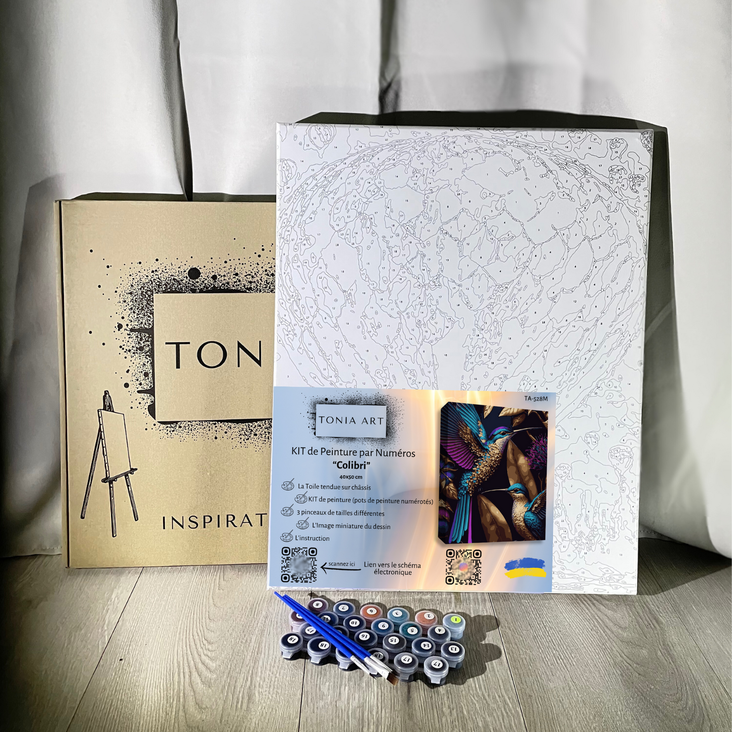 Kit de peinture par numéros de la marque Tonia Art, comprenant une toile imprimée avec le dessin d'une montgolfière, des pots de peinture numérotés, trois pinceaux de différentes tailles, et une boîte en carton marron avec le logo de la marque. Une petite image de référence colorée montre le résultat final.