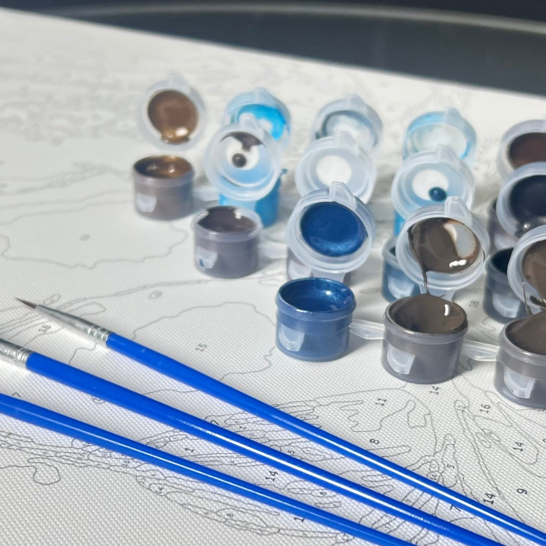 Ensemble de pots de peinture ouverts dans des tons bleus, gris et bruns disposés sur une toile de peinture par numéros. Trois pinceaux bleus sont posés au premier plan, prêts pour la réalisation du tableau.