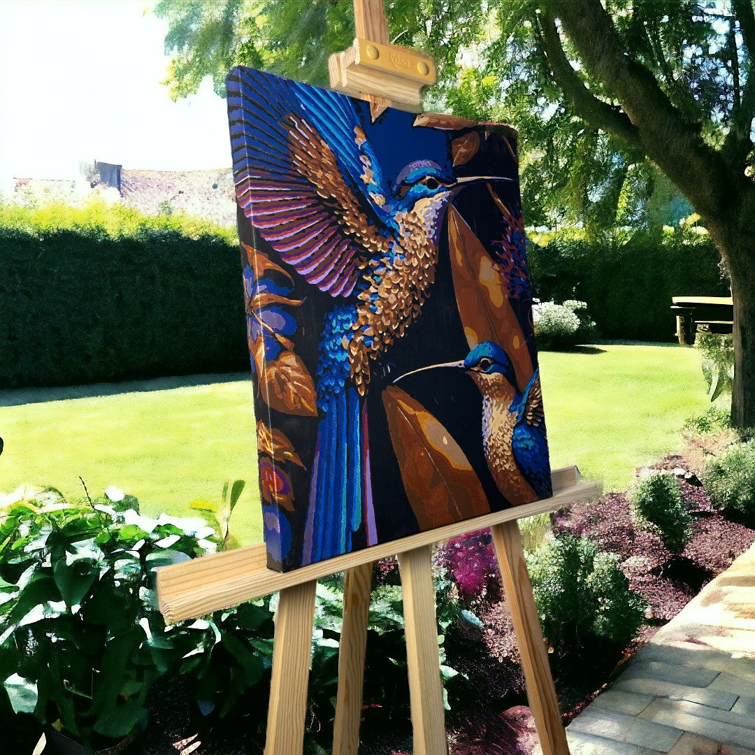Peinture de colibris aux couleurs vives, montrant deux oiseaux aux plumes bleues et dorées, posée sur un chevalet en bois dans un jardin verdoyant. Le décor extérieur inclut une pelouse, des buissons et des arbres, baignant l'œuvre dans la lumière naturelle.