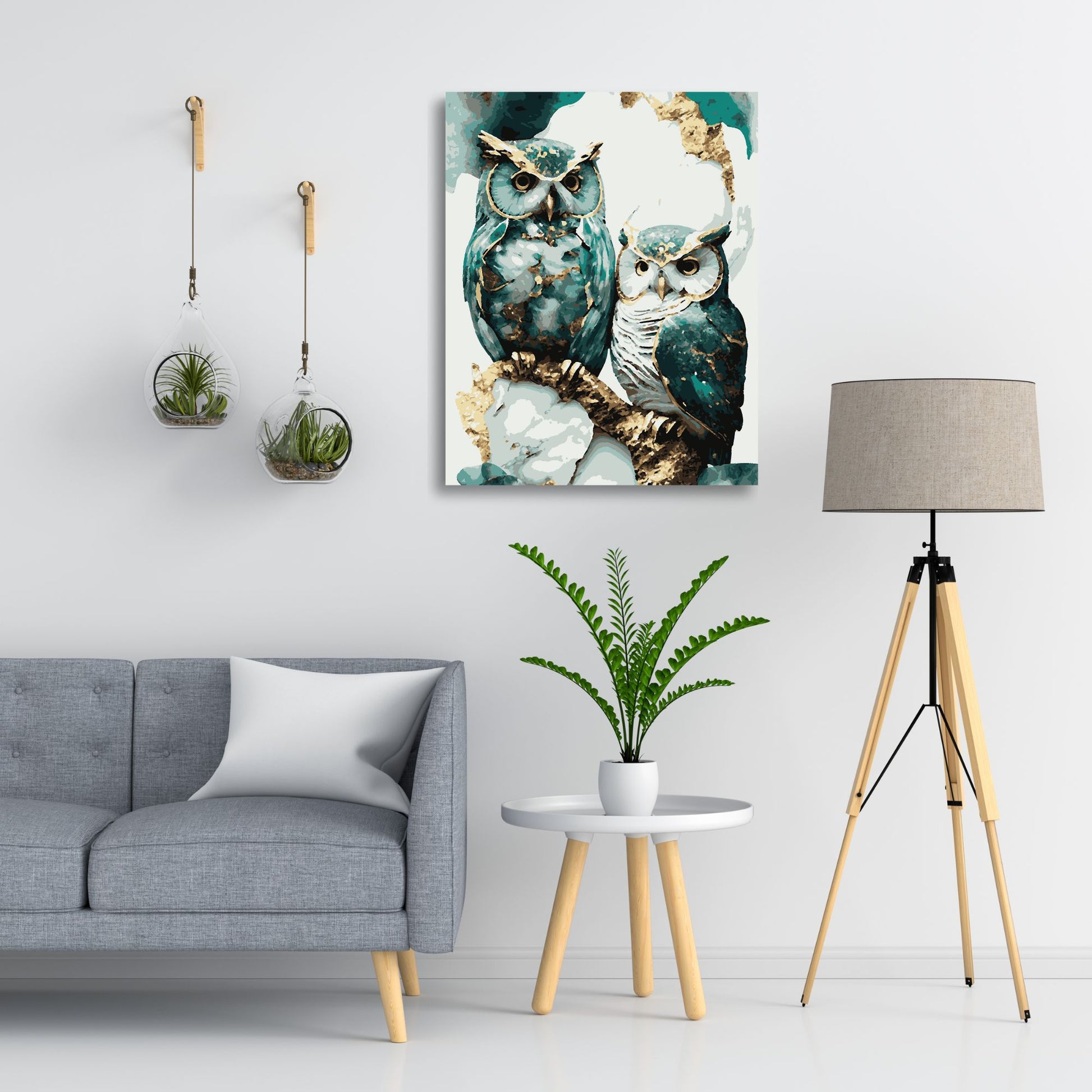 Intérieur moderne avec un canapé gris, une table d'appoint blanche avec une plante en pot, une lampe sur trépied en bois et une peinture d'un duo de hiboux aux plumes bleu-vert et dorées accrochée au mur. Deux terrariums suspendus en verre ajoutent une touche naturelle à l'espace